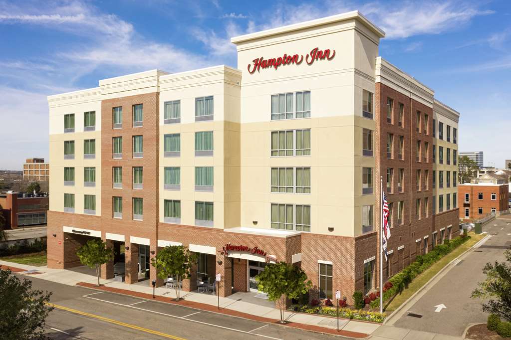 Exterior view of a five‑story Hampton Inn hotel with red-brick and cream-colored façade, the ‘Hampton Inn’ logo above the entrance, and an American flag flying on a pole out front.